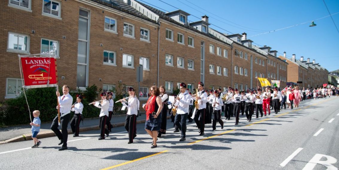 Korpset er ute og marsjerar  på Landås. Her ser me fana og heile korpset bak, før skulen følgjer etter.