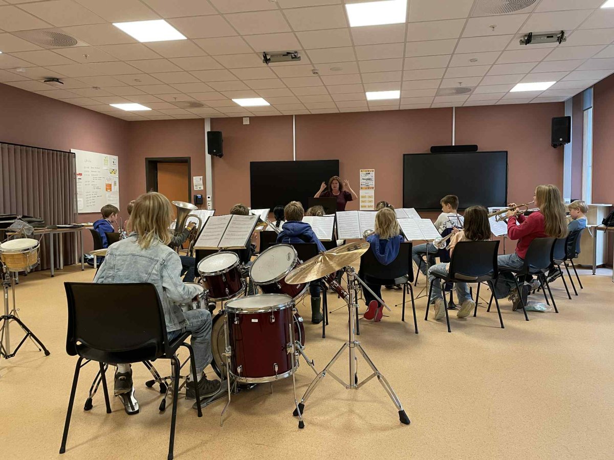 Juniorkorpset bakfrå. Det er barn som spelar, trommesettet er nærast i biletet. Dirigenten står med ansiktet vendt mot fotografen, og dirigerer.